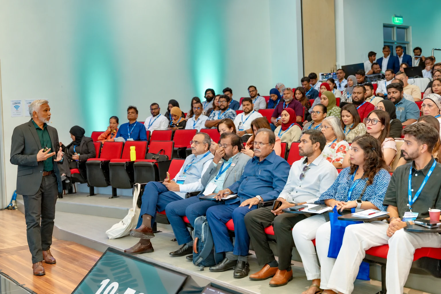 Mohamed Shareef standing at the front of a tiered auditorium, speaking with a full audience of conference participants