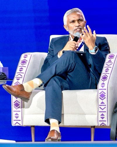Mohamed Shareef seated in an armchair on a blue stage, speaking into a microphone during a session