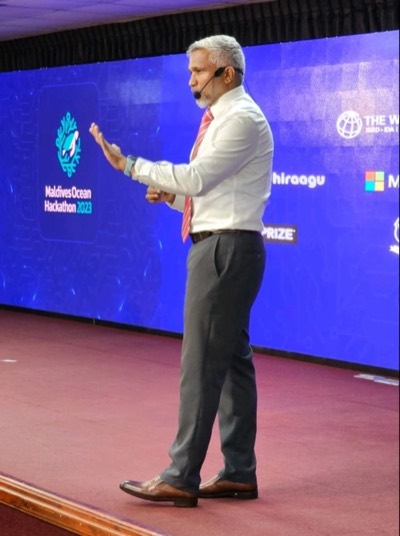 Mohamed Shareef on stage with a headset microphone, gesturing as he speaks, in front of a screen showing the Maldives Ocean Hackathon 2023 logo